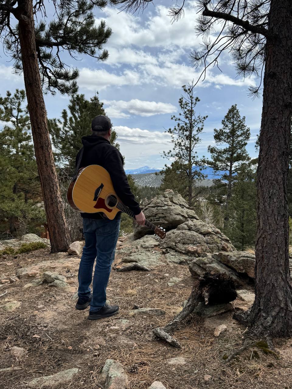 Lance holding his acoustic guitar among the pines, looking out at snowy mountain peaks